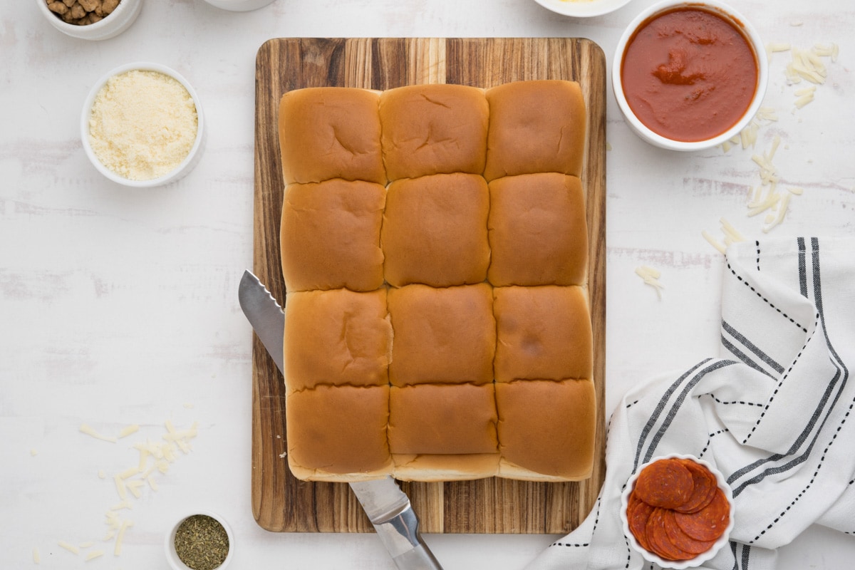 Slab of hawaiian rolls with a serrated knife on a wooden cutting board.