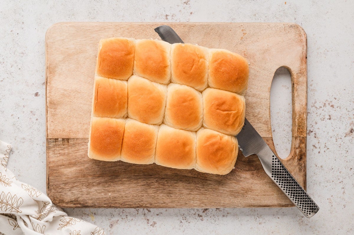 Hawaiian rolls on a wooden cutting board with a serrated knife slicing through them.