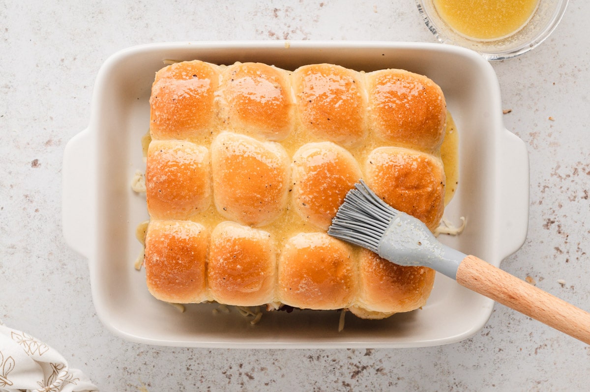 A gray pastry brush, brushing butter on top of a slab of slider rolls in a baking dish.