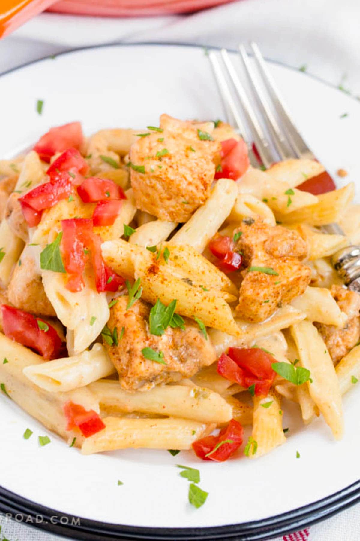 White plate with pasta and chicken with diced tomatoes and a creamy sauce and a fork.