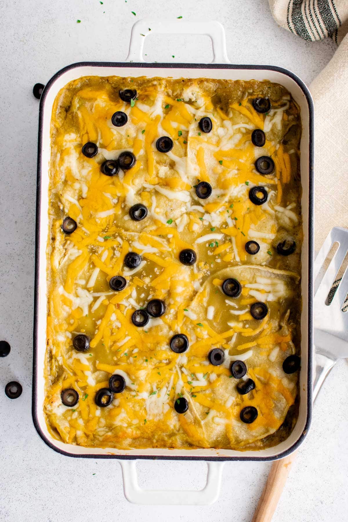 Casserole dish with green sauce, shredded cheese and olives.