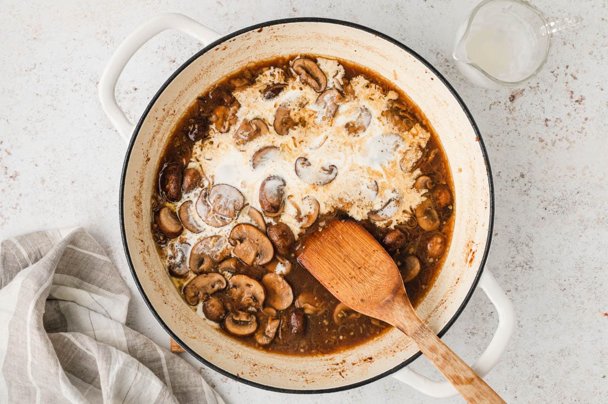 Mushrooms and a brown sauce with cream in a large pan with a wooden spatula.