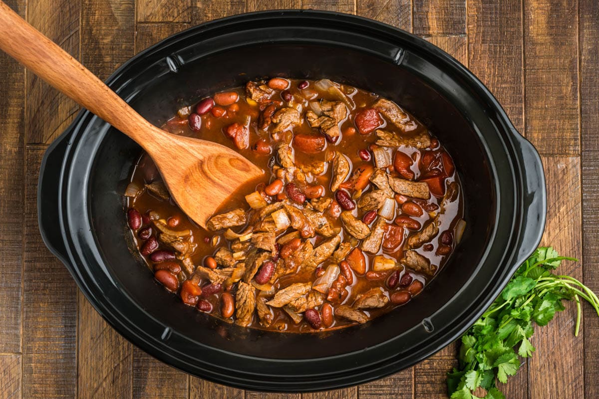 Steak chili in a slow cooker with a wooden spoon.