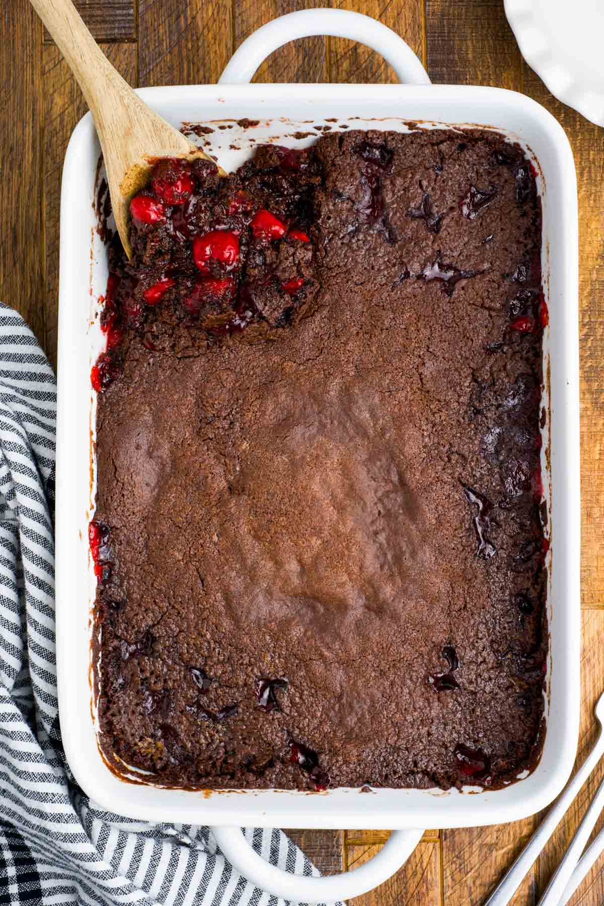 Wood spoon scooping chocolate cherry cake from a white baking dish.