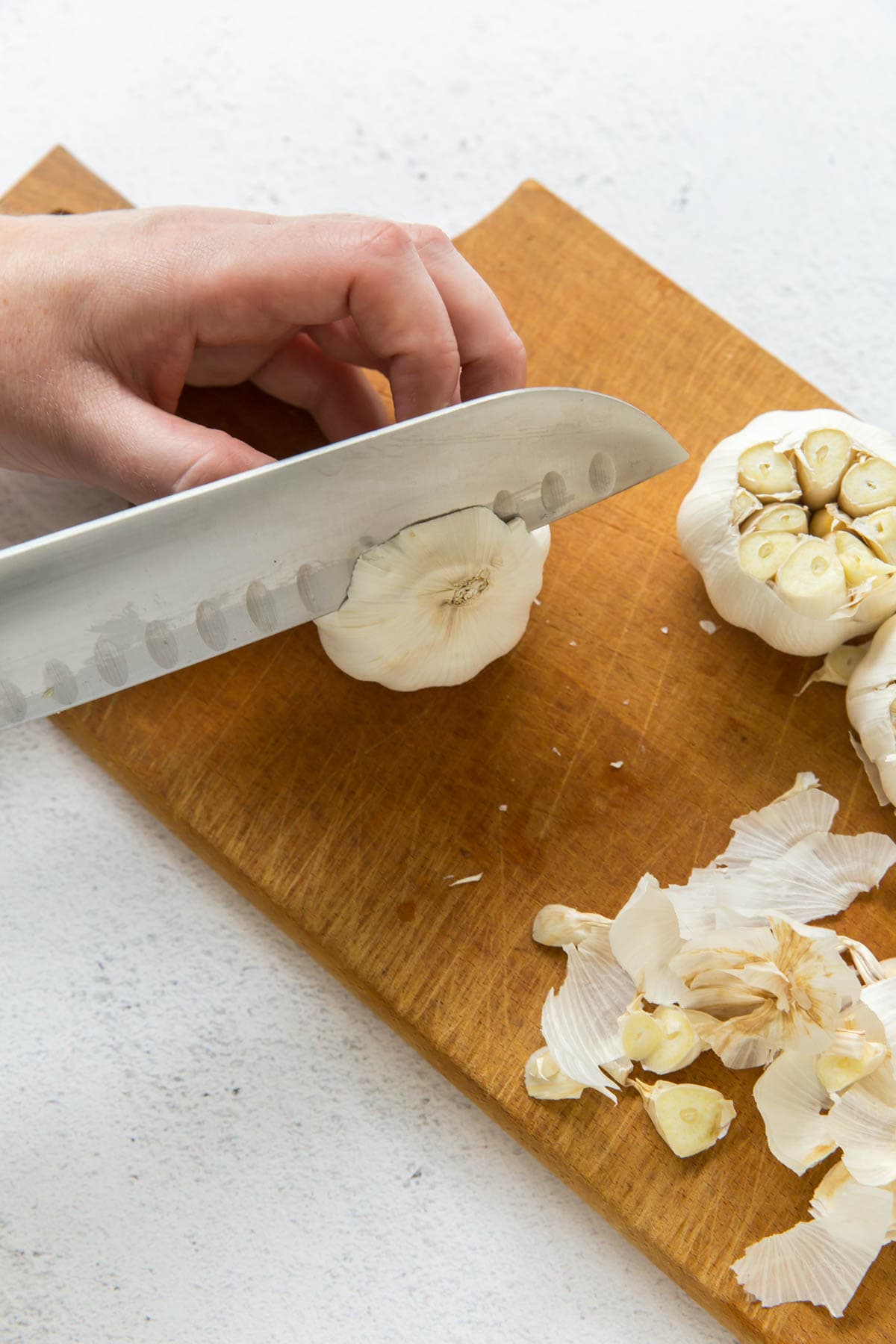 A wood cutting board, head of garlic, and a hand holding the bulb and a knife slicing one end of the garlic off.