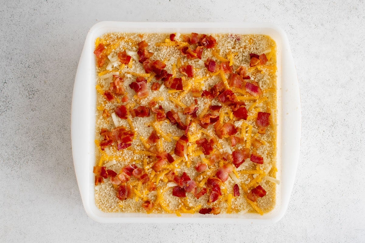 Mac and cheese, bacon and shredded cheese in a square baking dish.