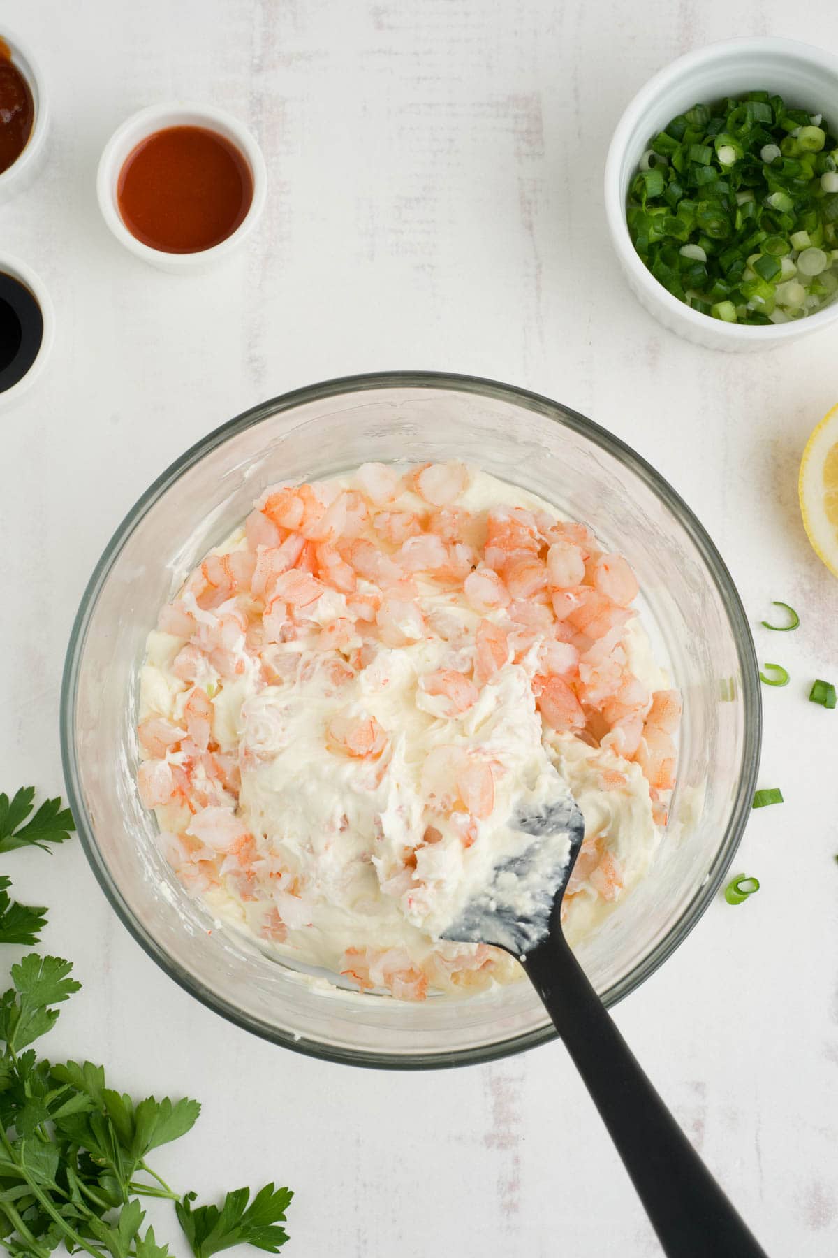 Chopped shrimp and cream cheese in a clear glass bowl with a black mixing spoon.