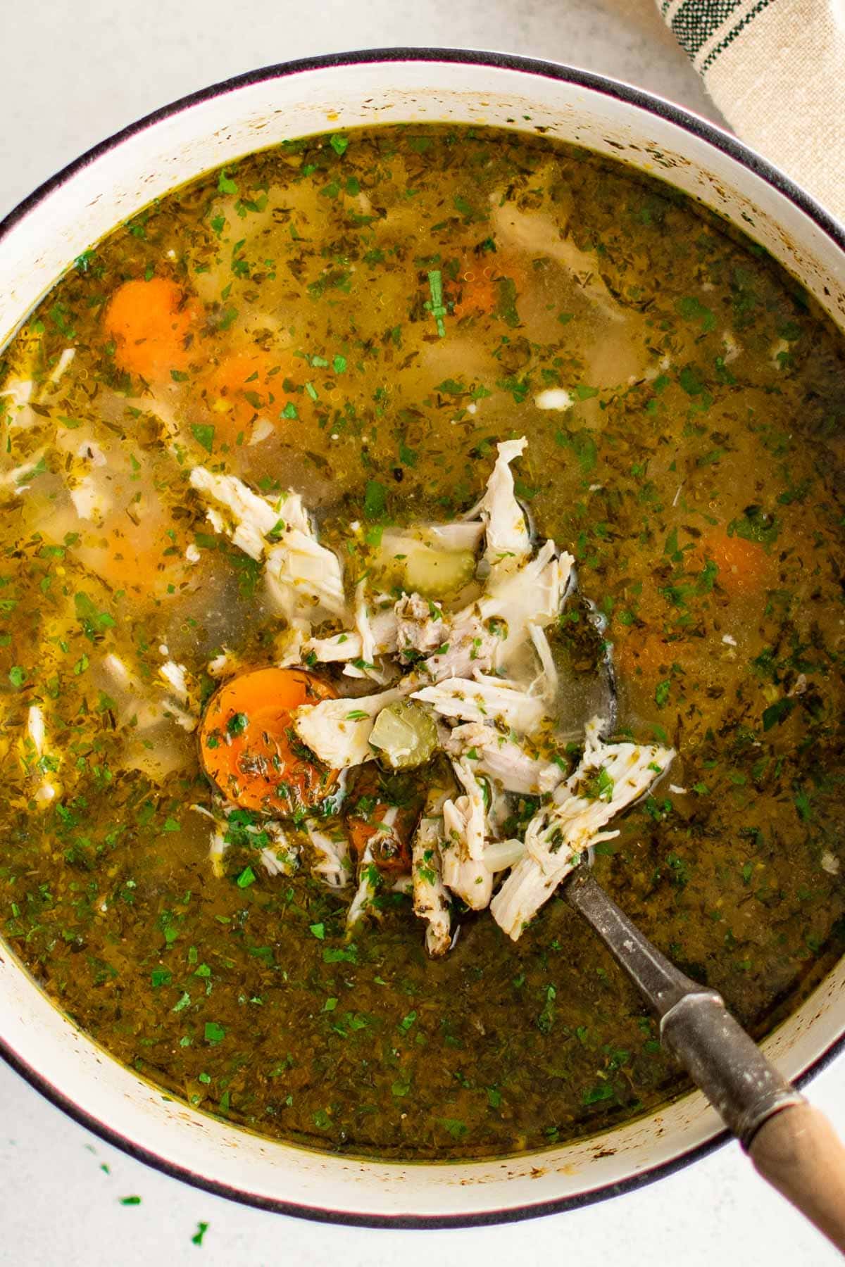 Shredded chicken, sliced carrots, herbs in chicken broth in a large soup pot, with a ladle.