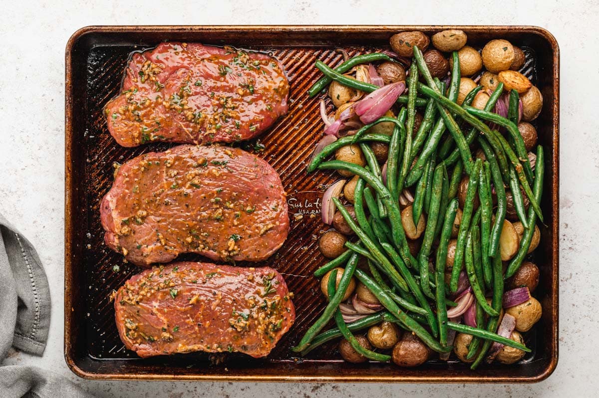 3 large steaks, green beans, potatoes and onions on a sheet pan.