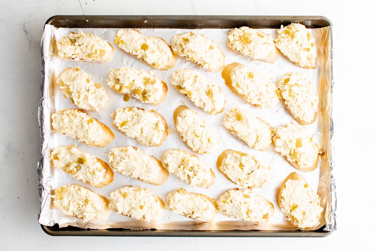 Sliced baguette on a baking sheet with butter and cheese mixture spread over each slice.