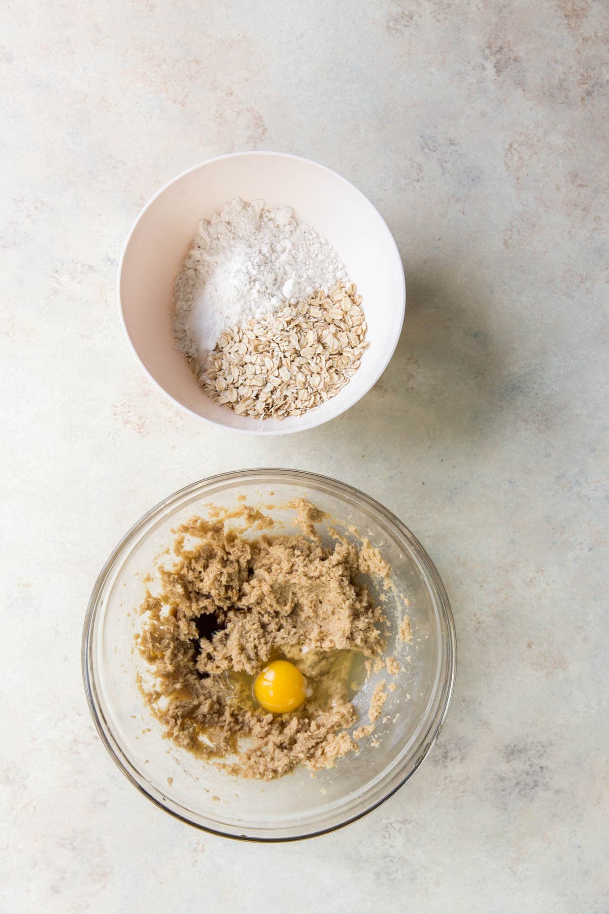 Two bowls, one with oats and flour, and the other with a light tan mixture and an egg.