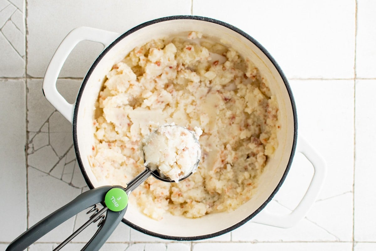 Mashed potatoes in a large pot, a cookie scoop holding some of the mashed potato.