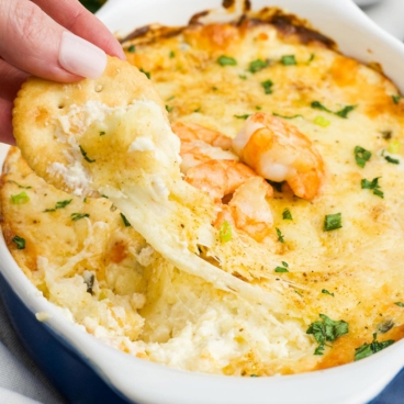 Baked shrimp dip with a Ritz cracker and a hand pulling a serving out.