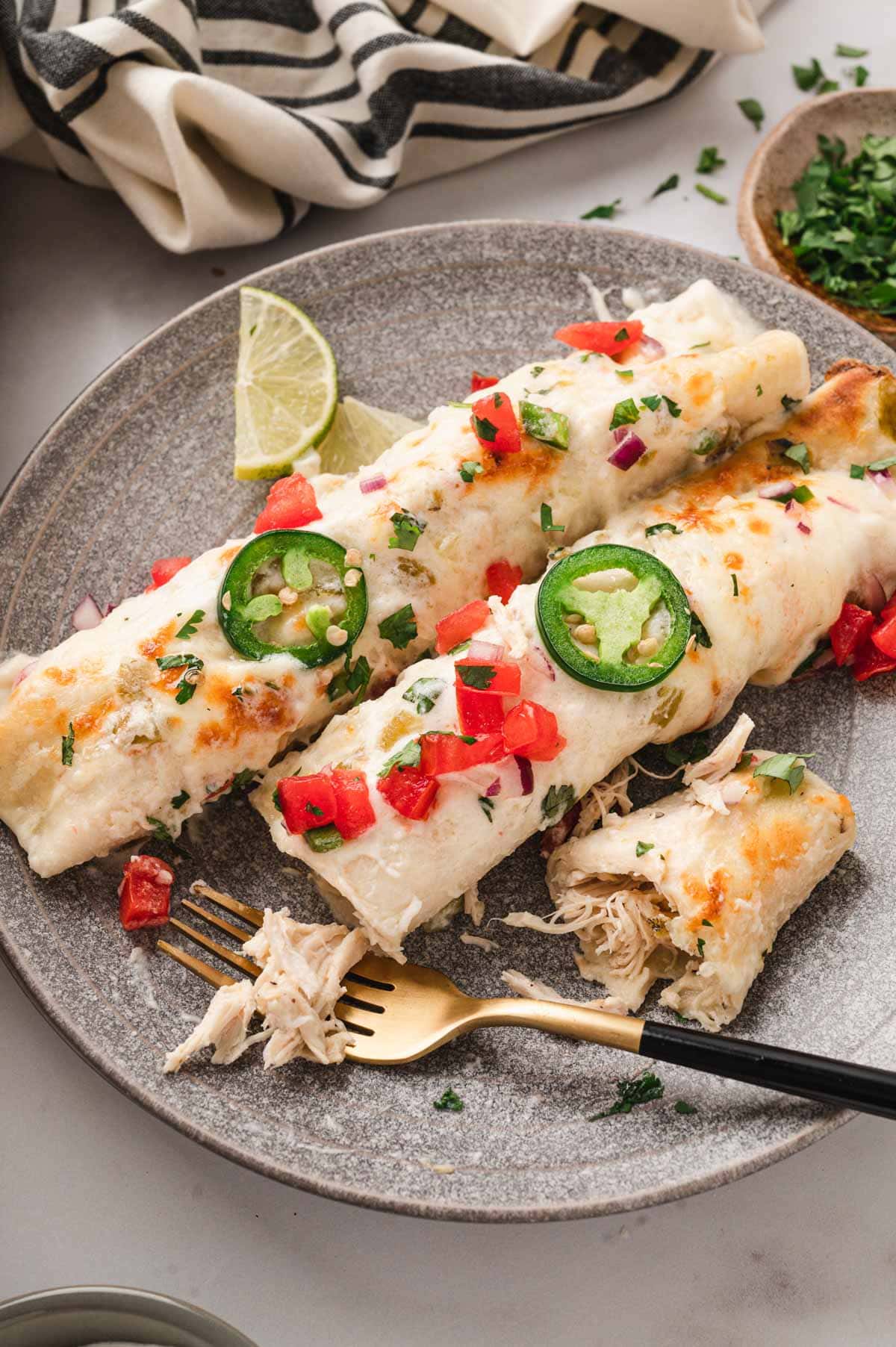 Two white enchiladas with diced tomato and sliced jalapeno and limes on a plate with a fork.