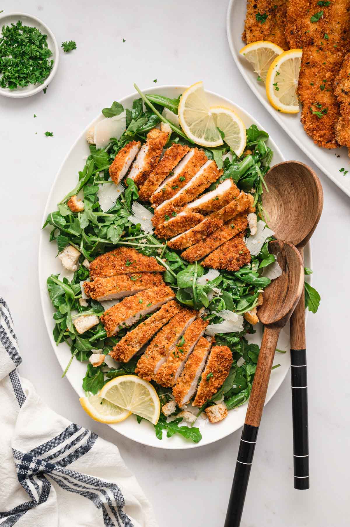 Crispy breaded chicken on a bed of arugula with lemon slices and two wooden serving spoons.