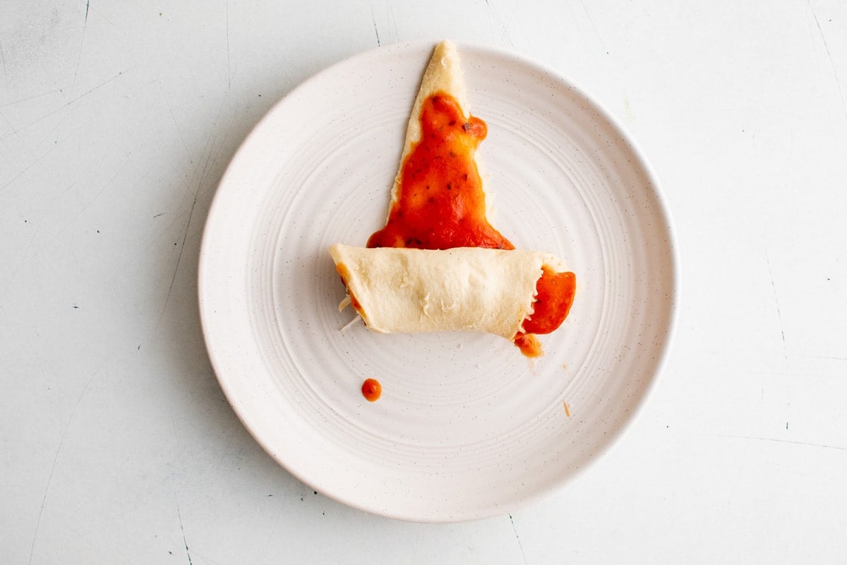 Crescent dough triangle with red sauce and pepperoni, being rolled up. On a white plate.