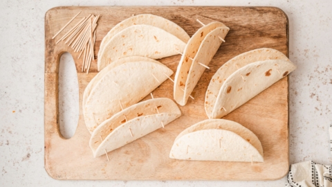 Flour tortillas folded in half with toothpicks in them, sitting on a wooden cutting board.