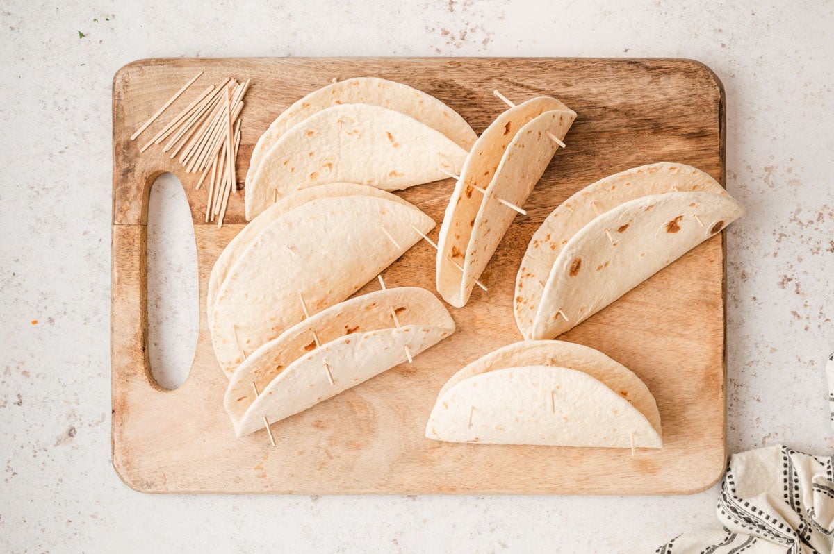 Flour tortillas folded in half with toothpicks in them, sitting on a wooden cutting board.