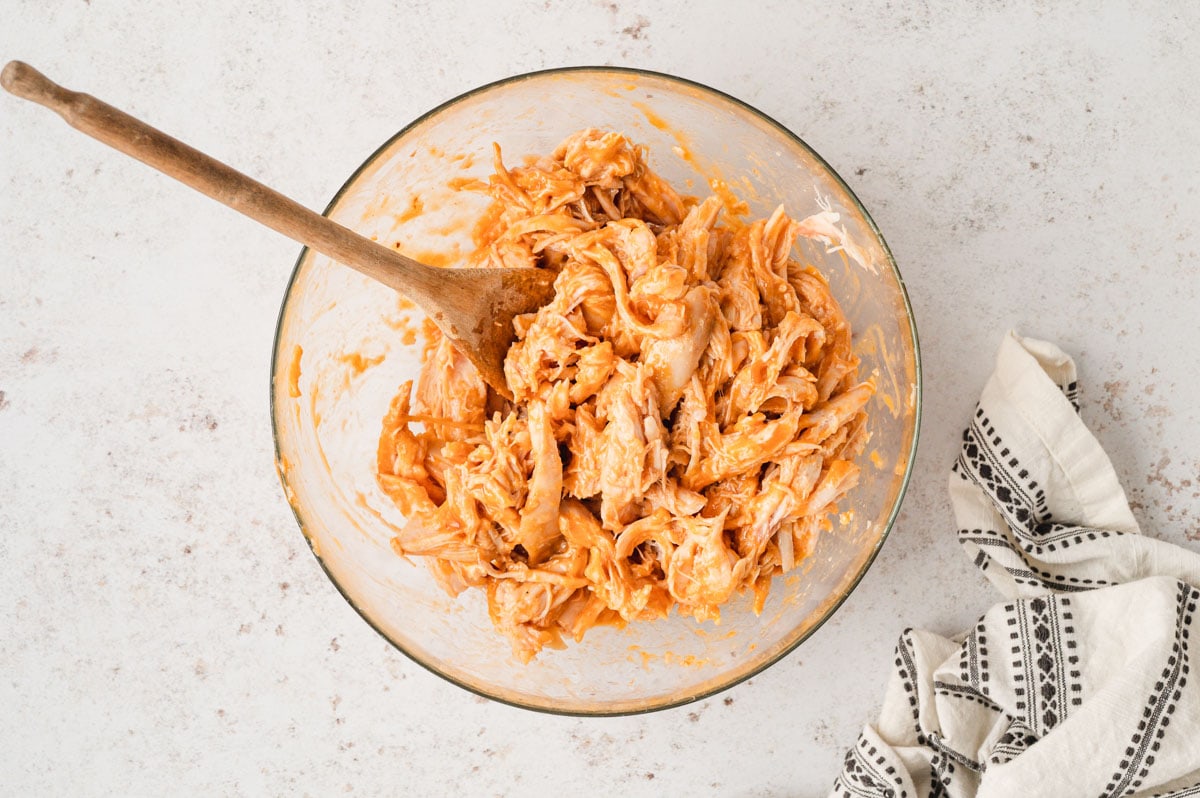 Buffalo shredded chicken in a large glass bowl with a wooden spoon.