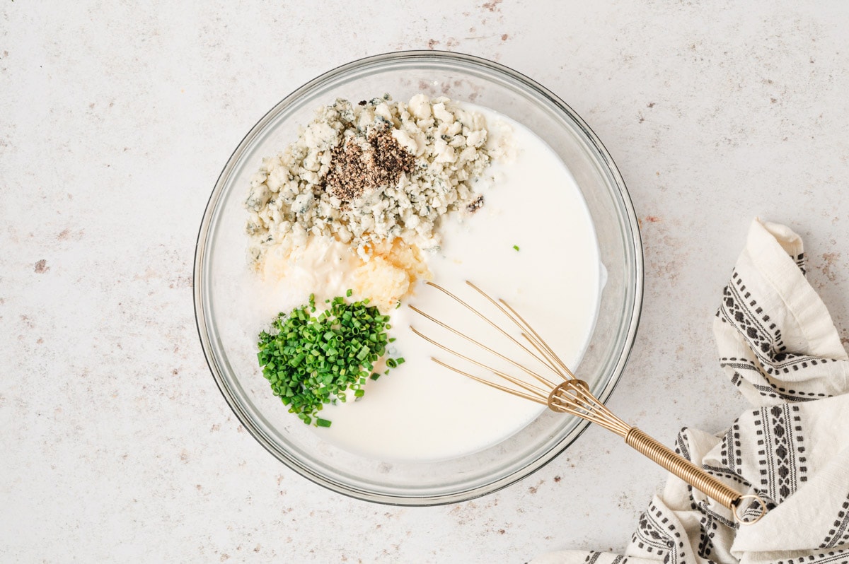 Buttermilk, blue cheese, pepper and chives in a large glass bowl with a whisk.