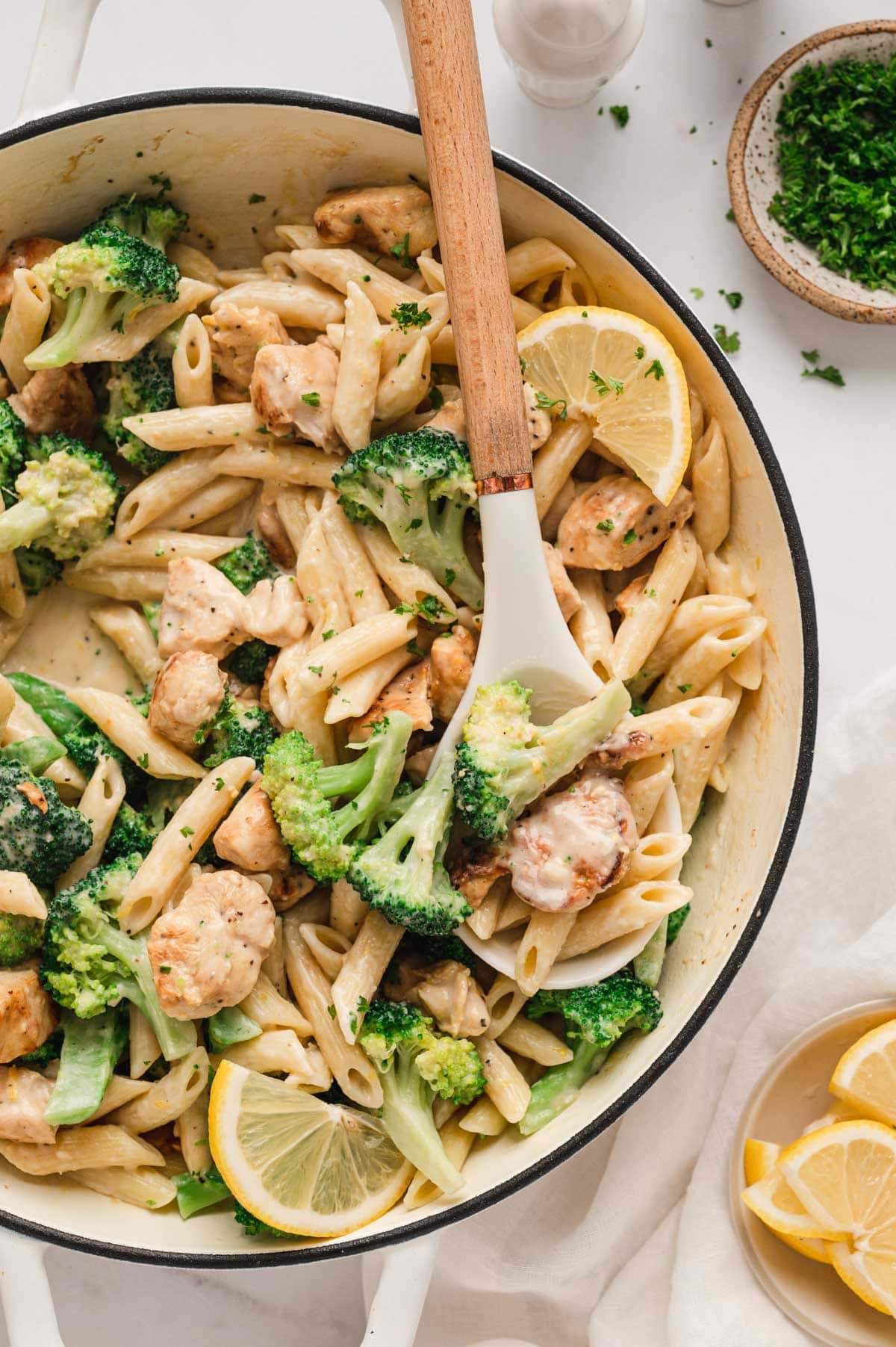 Pan of broccoli, chicken and pasta with lemons and a wooden spoon.