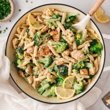 Chunks of chicken, broccoli, penne pasta and lemon slices in a skillet with a spoon.