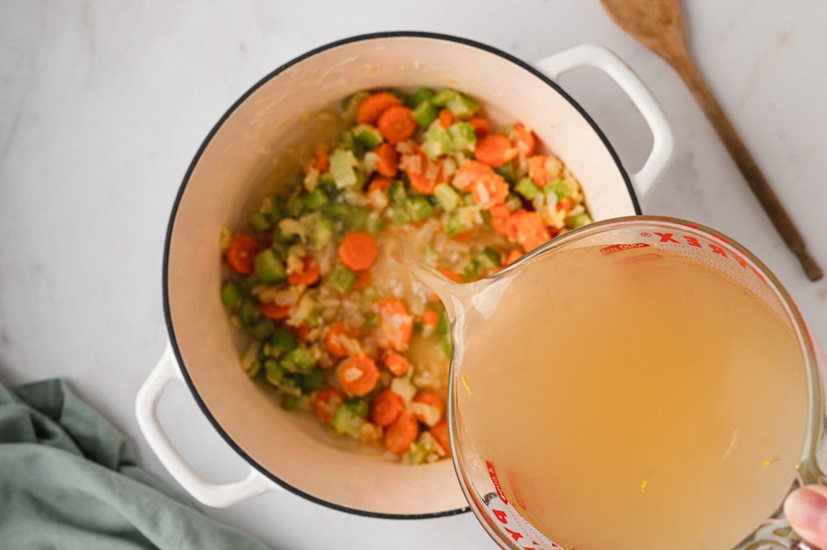 Broth pouring from a glass cup into a pot of carrots and celery.