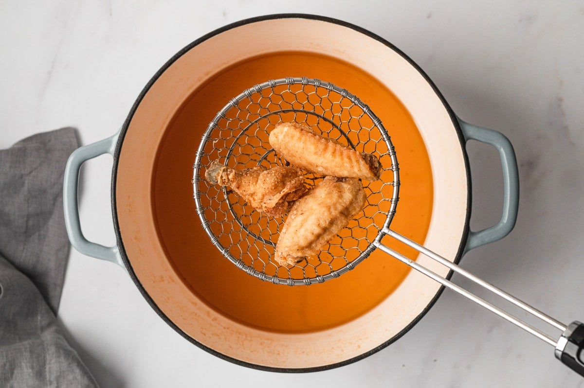 3 chicken wings in a wire mesh scoop held over a pot of oil.