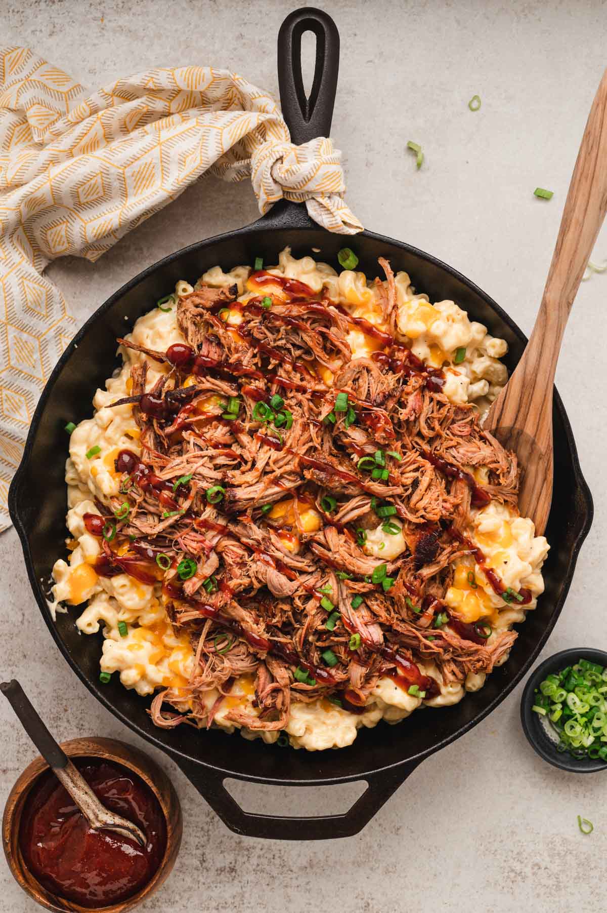Mac and cheese in a cast iron skillet topped with bbq pulled pork, and with a wooden spoon.