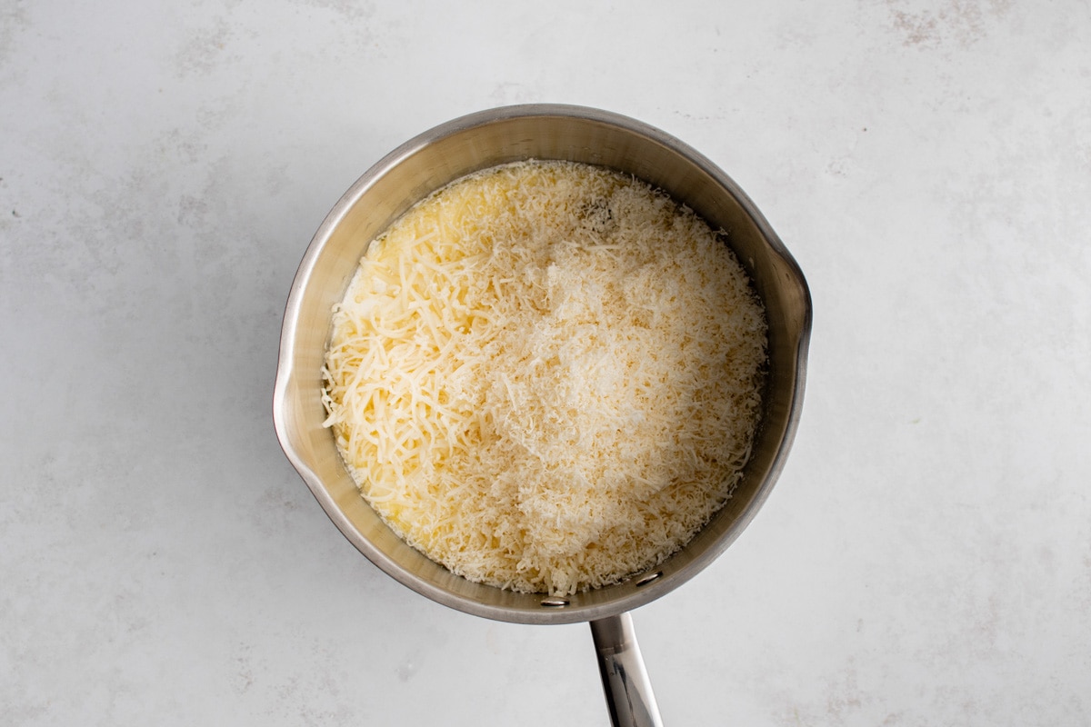 Shredded cheese on top of a white sauce in a metal saucepan.