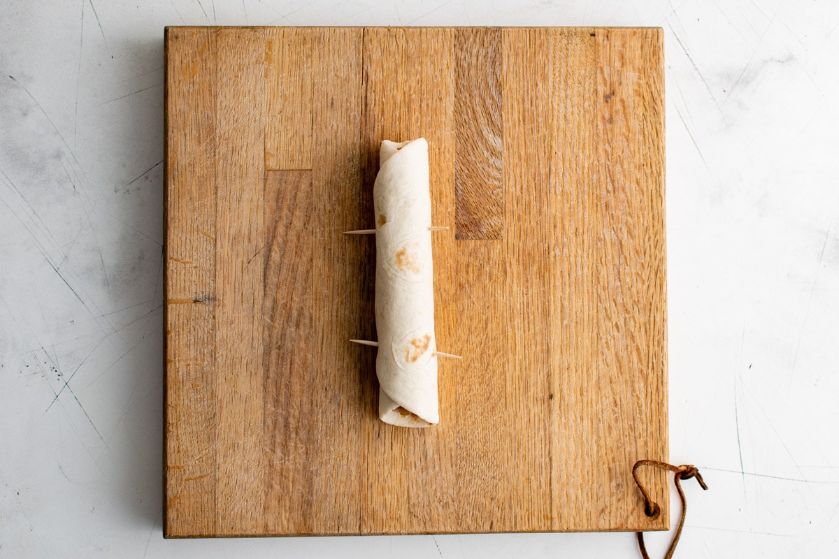 Rolled up flour tortilla with toothpicks on a wooden cutting board.