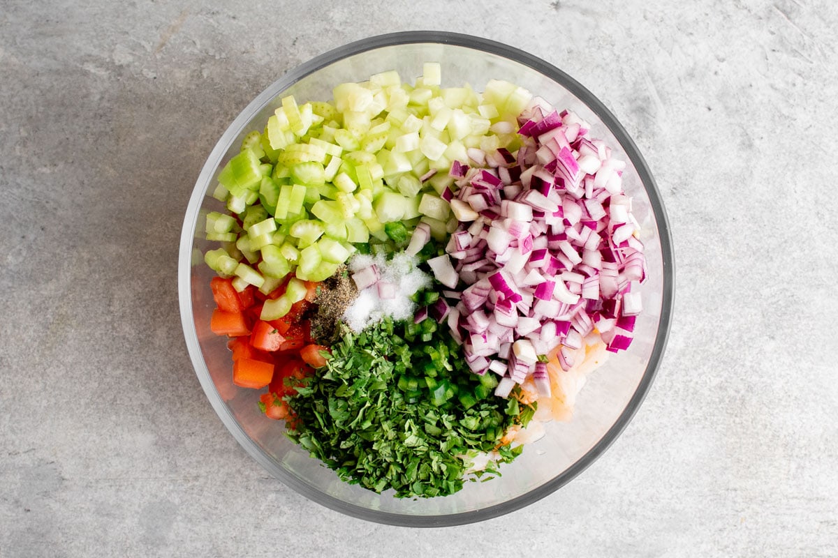 Diced red onions, celery, cucumber, cilantro, tomatoes in a clear glass bowl.