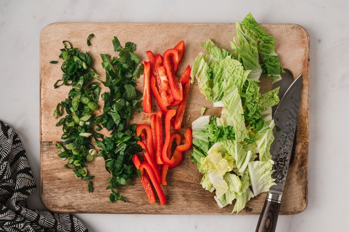 Chopped cabbage, red bell pepper, cilantro and green onions on a wooden cutting board.