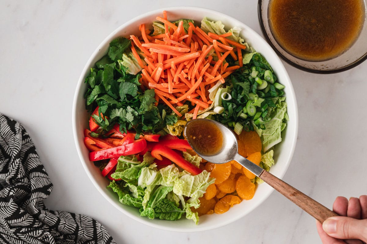 Shredded carrots, napa cabbage, red bell peppers and oranges in a white bowl, a spoon with salad dressing ready to drizzle over the salad.