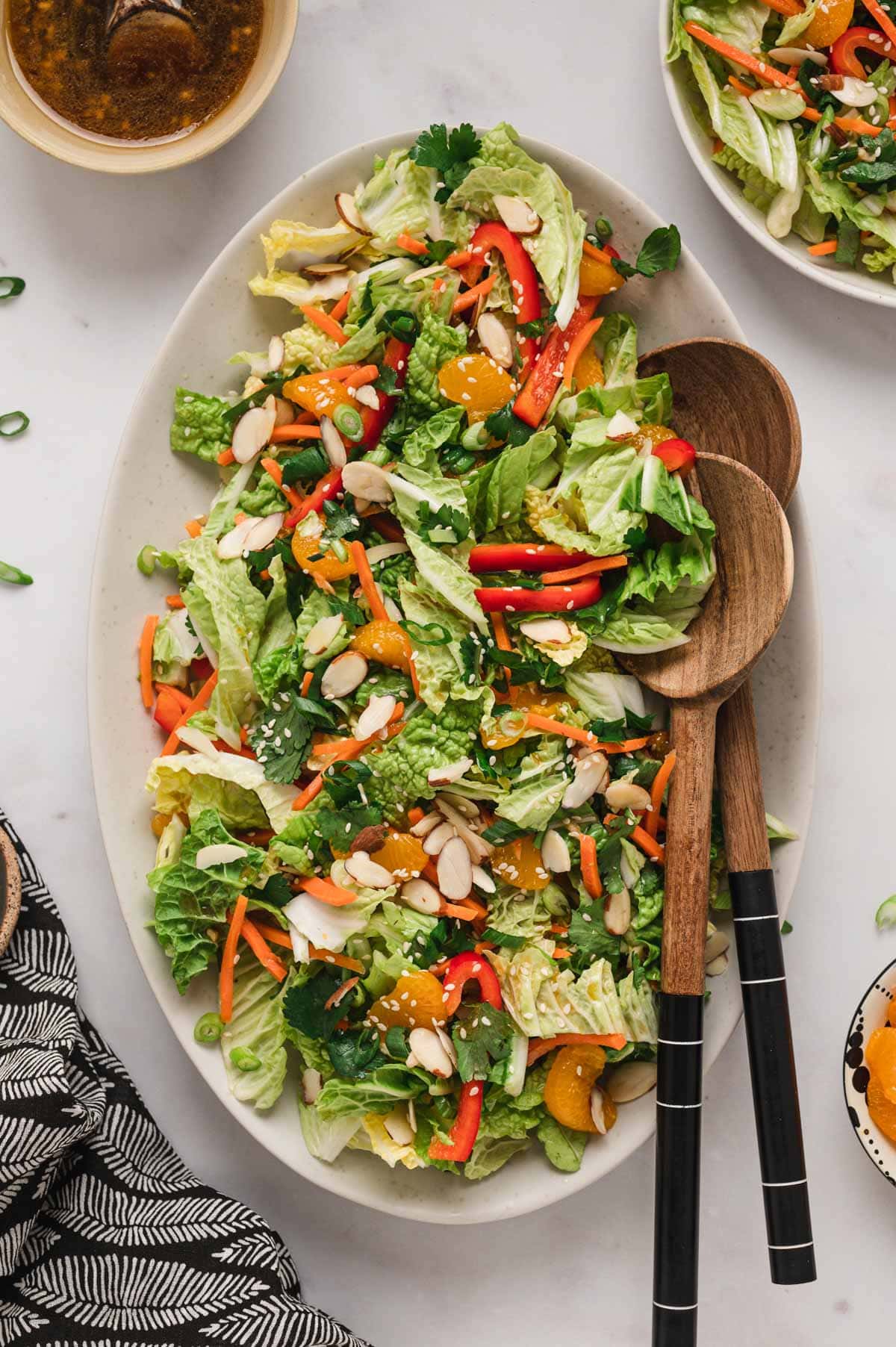 Salad with napa cabbage, red bell peppers and oranges on a white platter with wood salad tongs.
