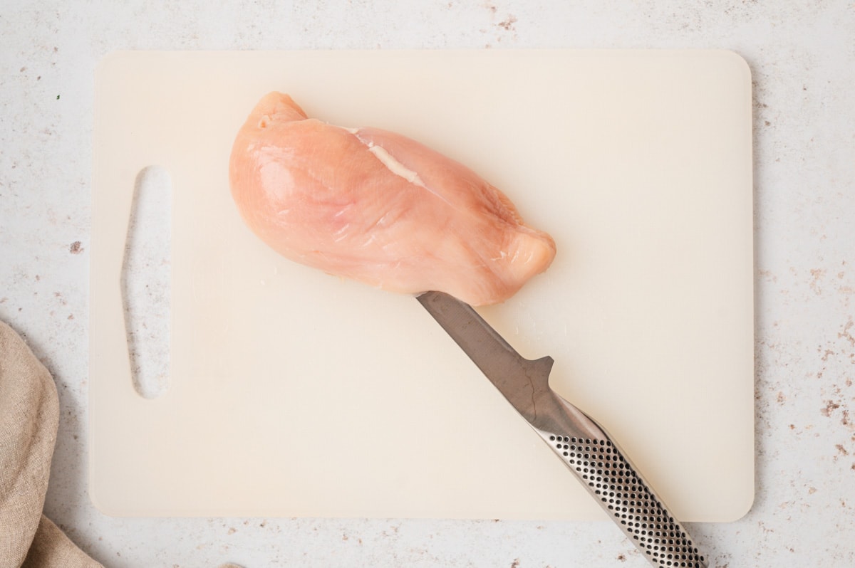 Uncooked chicken breast on a white cutting board, with a knife slicing through the center.