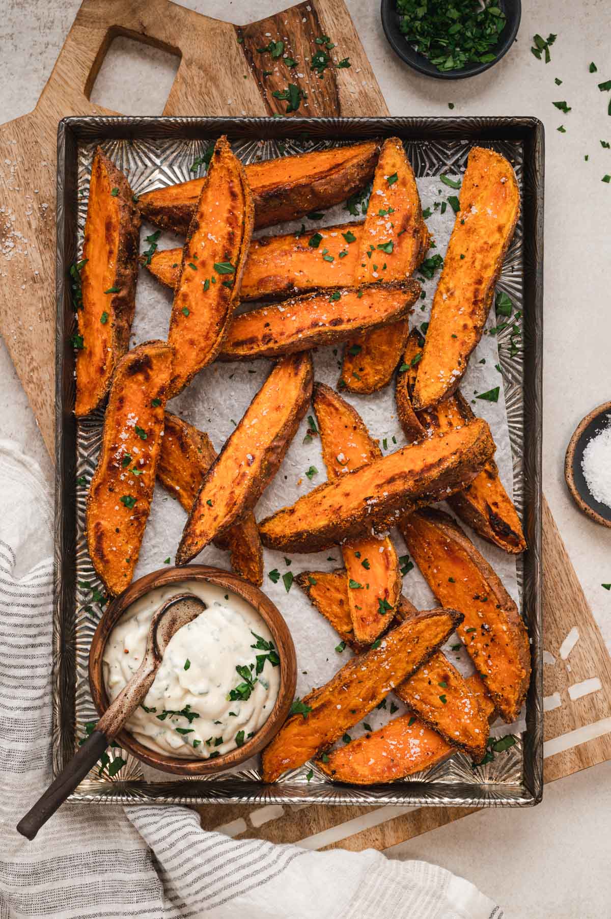 Sweet potato wedges on a baking sheet with minced parsley garnish and a creamy dipping sauce.