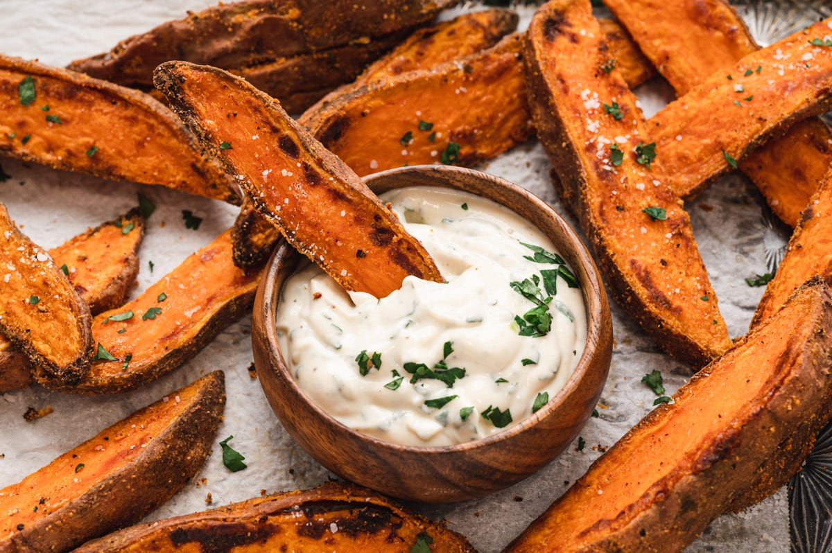 Sweet potato wedges and a wood dish of garlic aioli. One wedge is dipped in the aioli.