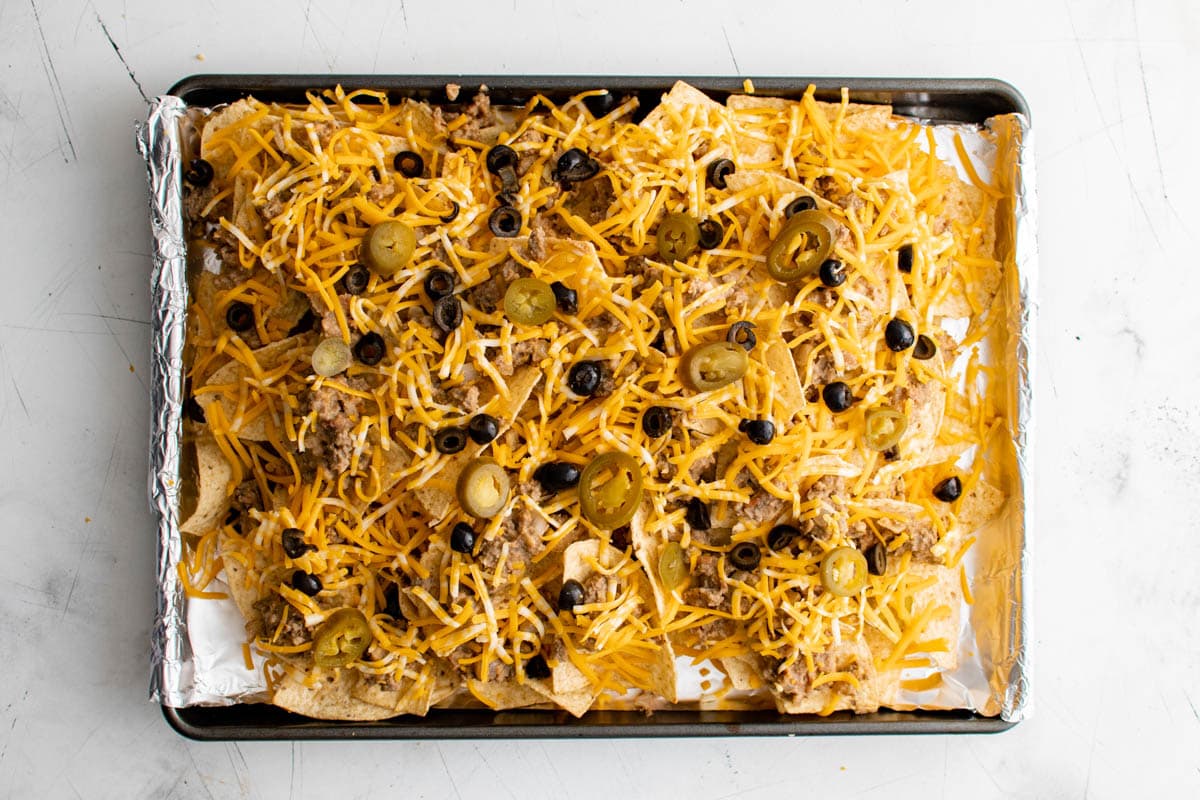 Nachos on a sheet pan with shredded cheese, sliced olives and sliced pickled jalapenos.