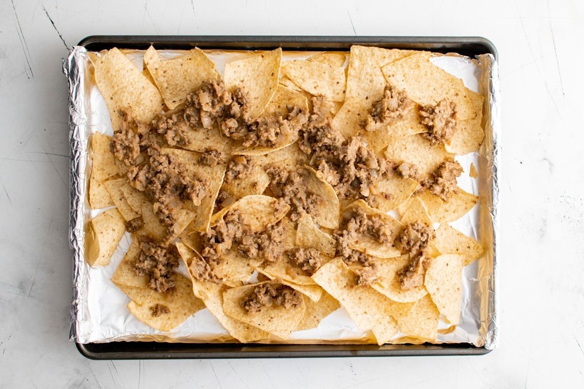 Tortilla chips on a foil lined sheet pan, topped with refried beans and ground beef.