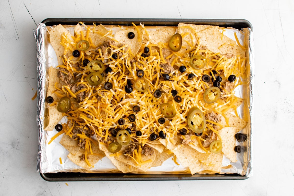 Tortilla chips on a lined sheet pan with shredded cheese and olives.