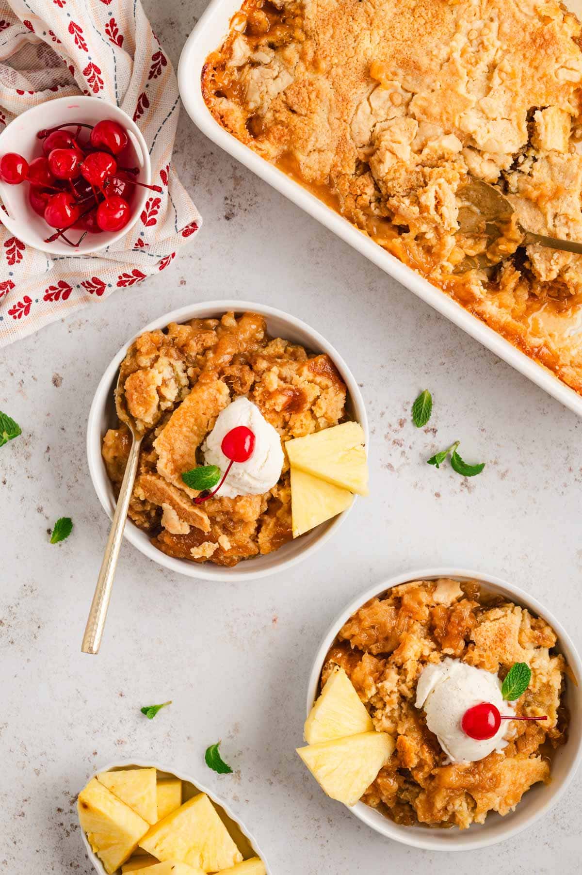 Pineapple dump cake, two serving bowls, with vanilla ice cream, sliced pineapple and cherries on top.