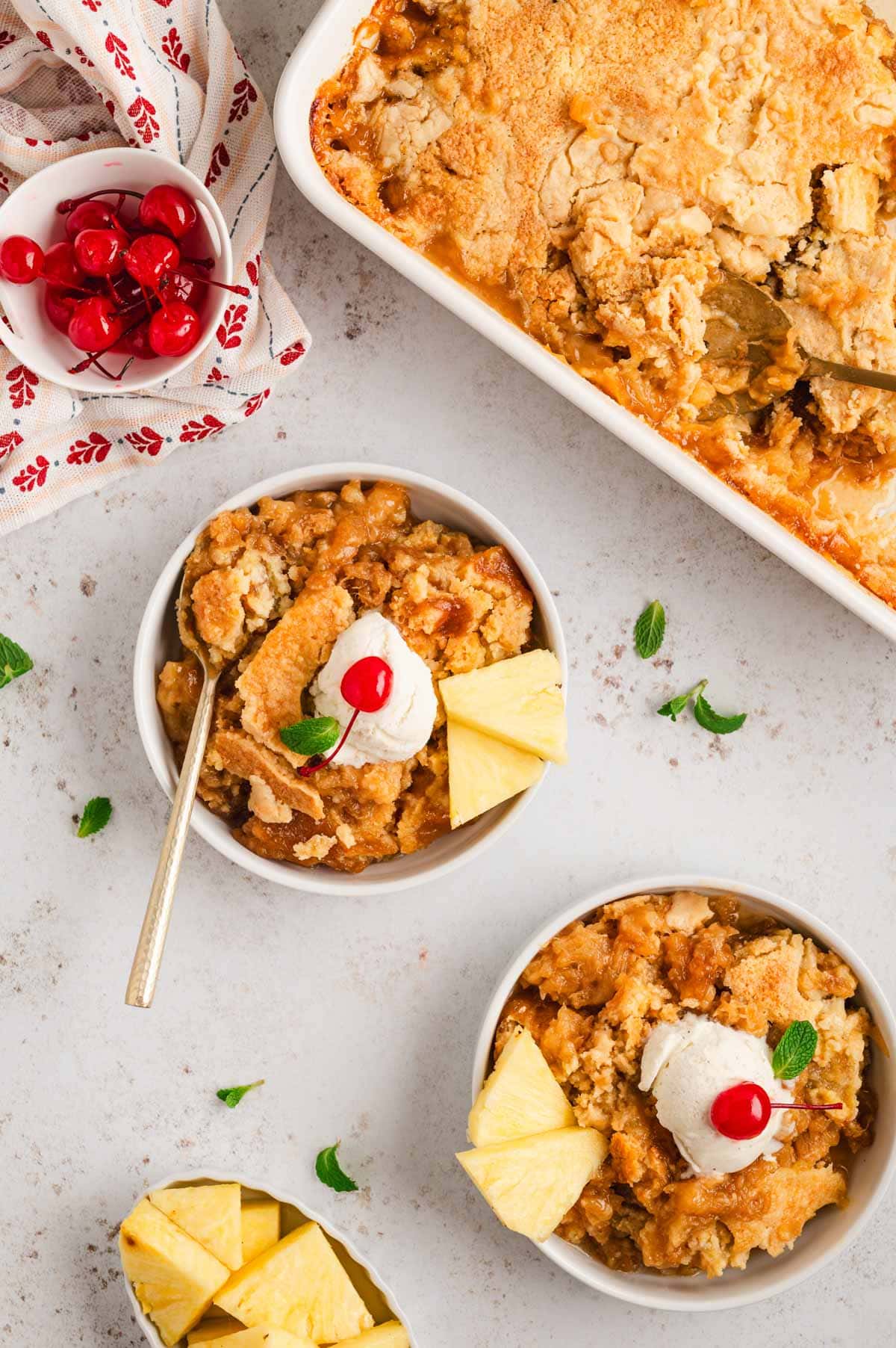 Pineapple dump cake, two serving bowls, with vanilla ice cream, sliced pineapple and cherries on top.