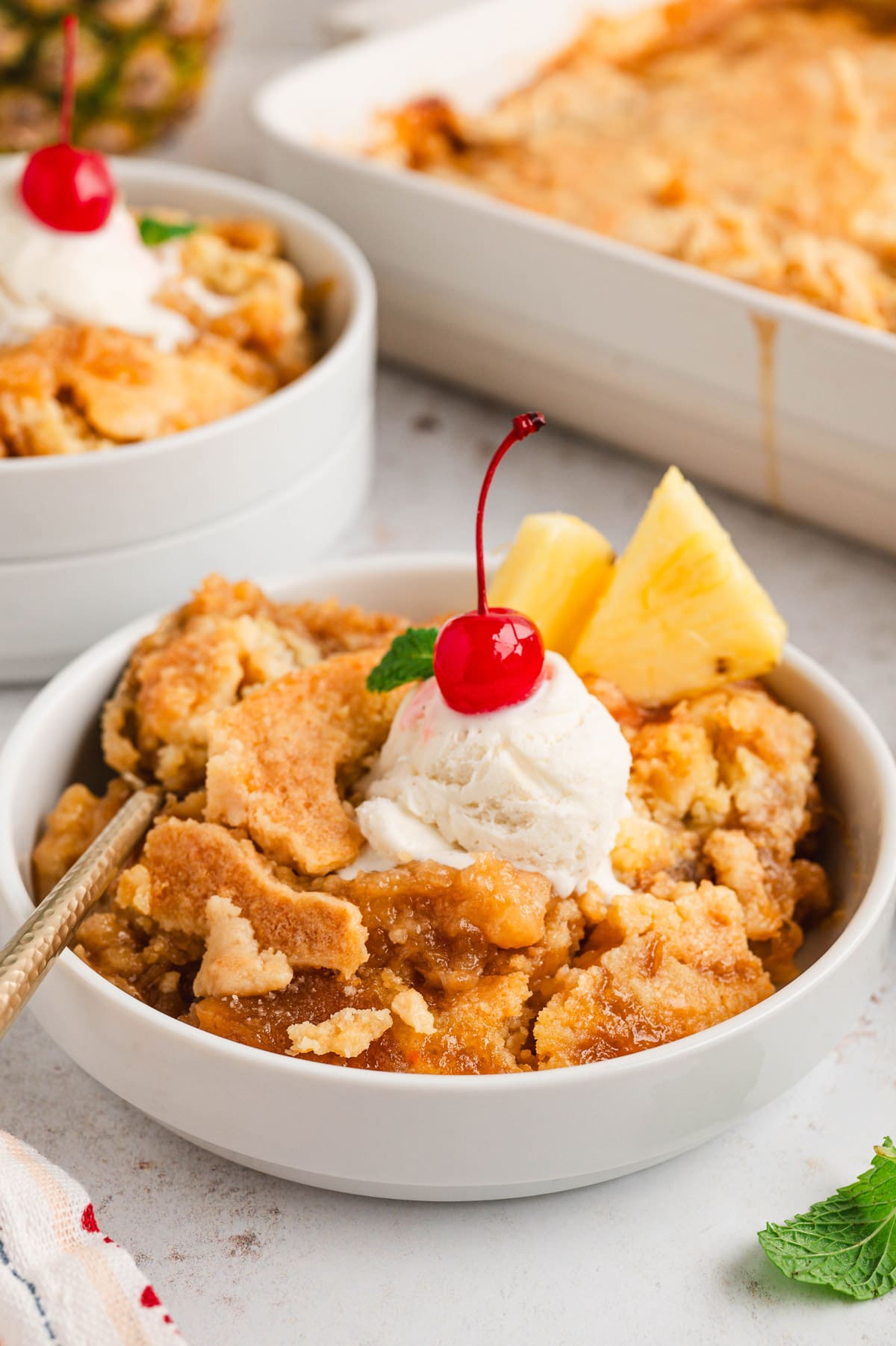 A serving of pineapple dump cake in a white bowl, with a scoop of vanilla ice cream, a cherry and fresh pineapple slices.
