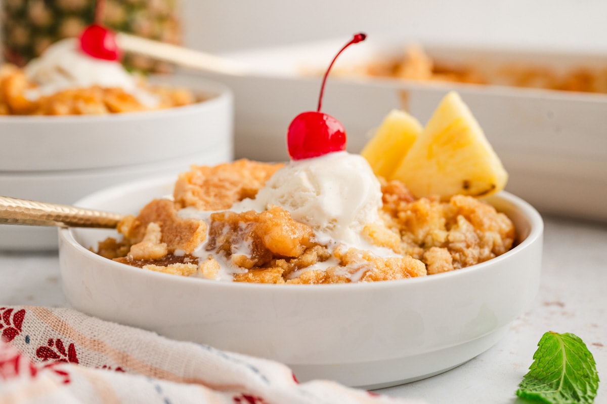 Pineapple dump cake in a white bowl with vanilla ice cream and a cherry on top.