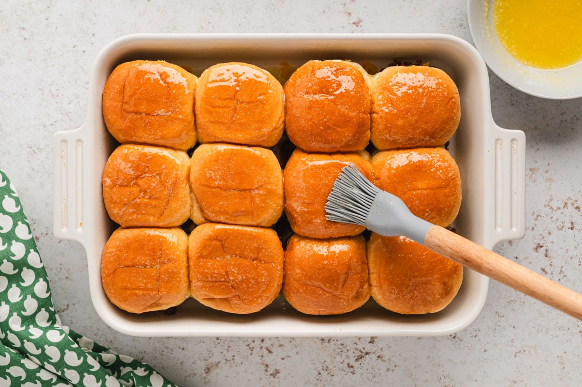 Slider rolls in a white baking dish brushed with melted butter and a gray silicone basting brush.