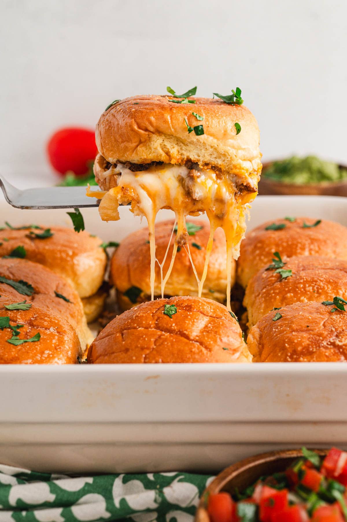 Sliders in a baking dish, a spatula lifting one out to reveal gooey melted cheese.