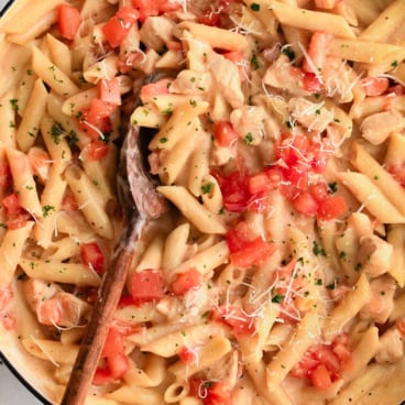 Square image of chicken and pasta witih tomaotes in a skillet.