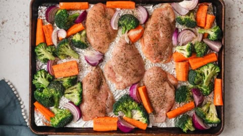 Uncooked chicken and broccoli and carrots on a sheet pan.
