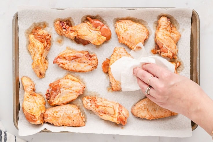 chicken wings on a baking tray with a hand patting dry with paper towel.